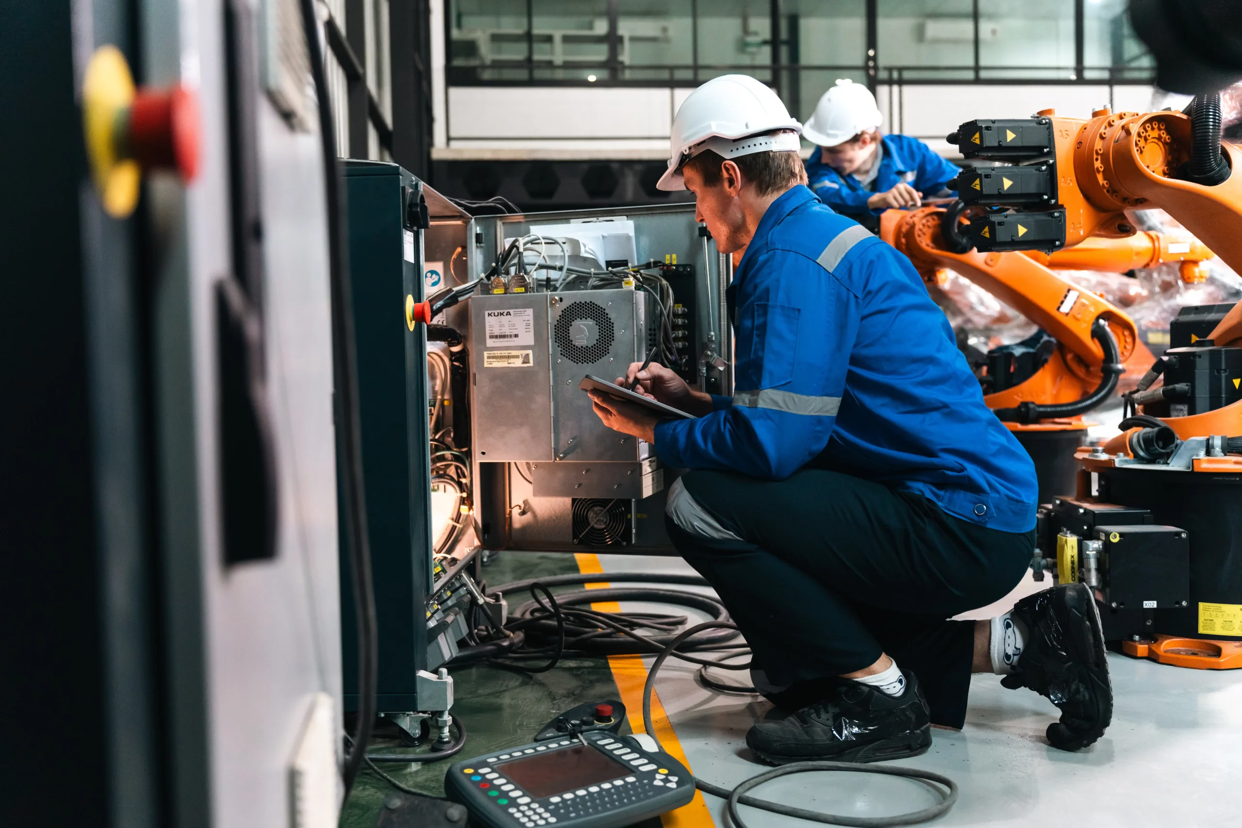 Técnico realiza manutenção em equipamento industrial automatizado, com auxílio de um tablet, enquanto outro profissional trabalha próximo a braços robóticos.