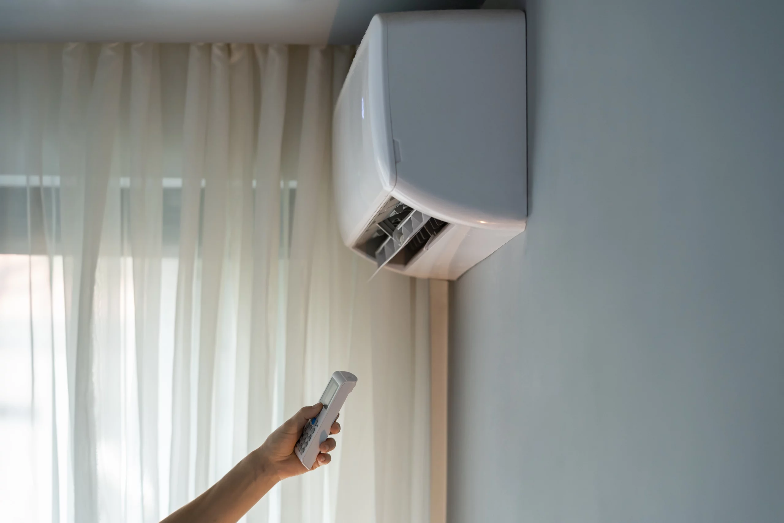 Pessoa usando controle remoto para ligar o ar-condicionado split em um quarto com cortinas claras.