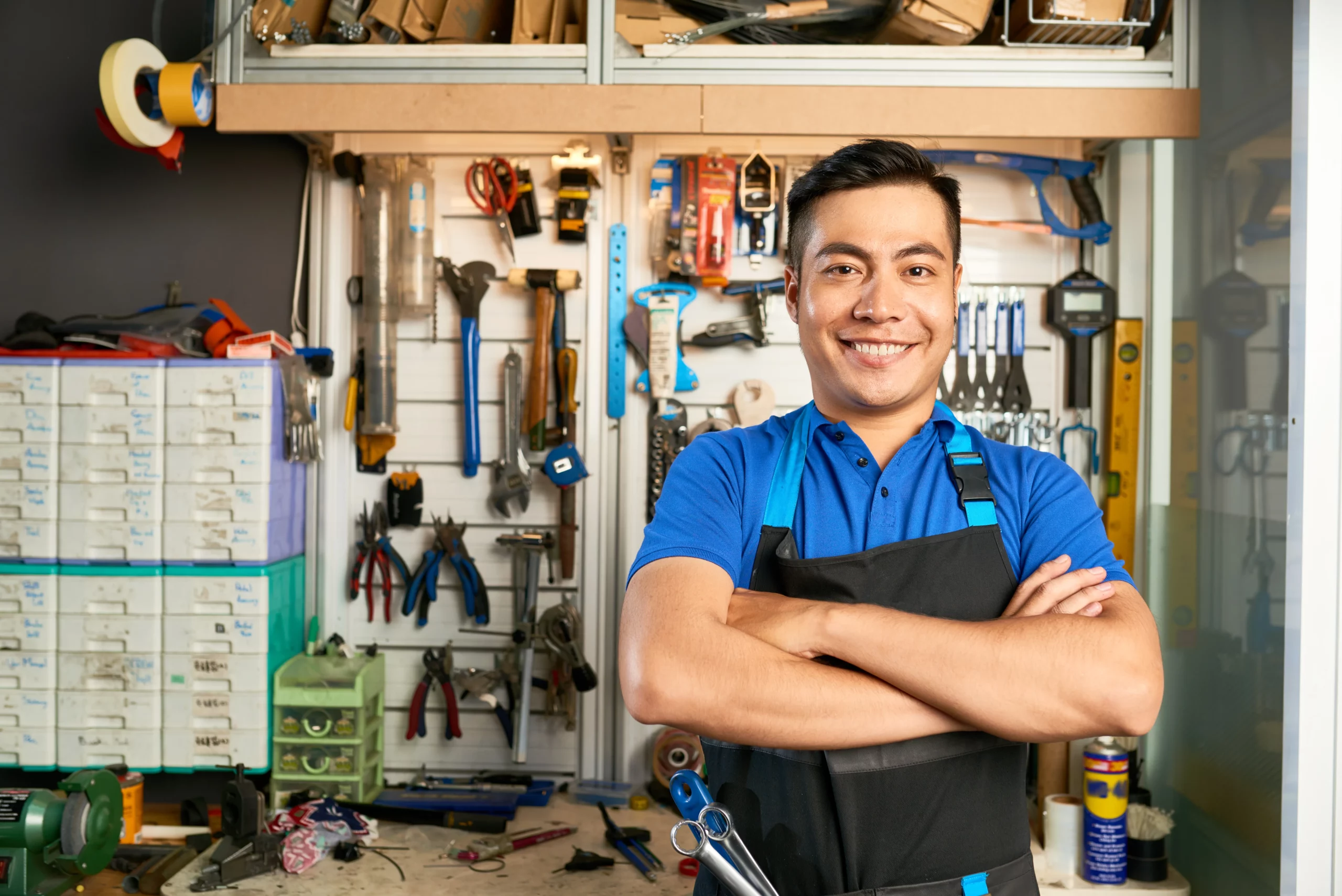 Marido de aluguel sorrindo, de braços cruzados, em uma oficina organizada com ferramentas penduradas em painel, bancada de trabalho e materiais de manutenção ao fundo.