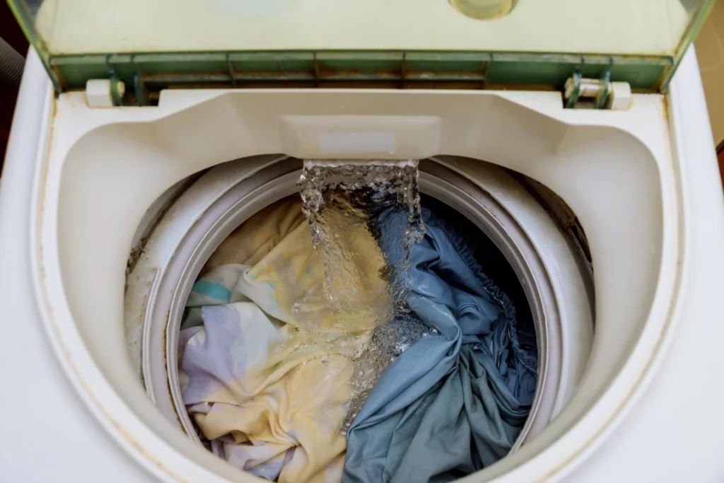 Vista interna de uma máquina de lavar aberta, com água corrente caindo sobre roupas coloridas dentro do tambor.
