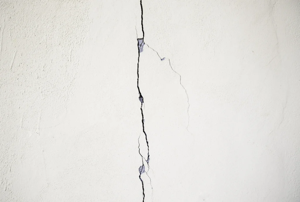 Rachadura na parede branca com fissura vertical visível e sinais de desgaste no revestimento, indicando possíveis problemas na estrutura ou acabamento.