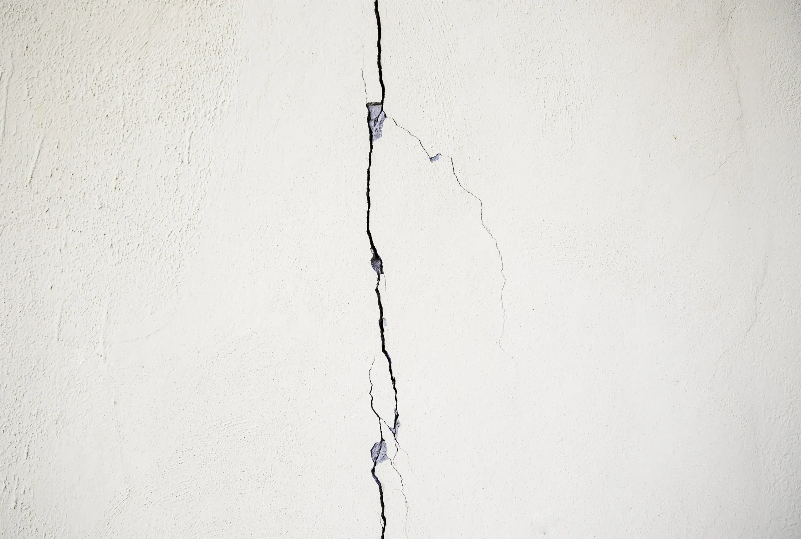 Rachadura na parede branca com fissura vertical visível e sinais de desgaste no revestimento, indicando possíveis problemas na estrutura ou acabamento.