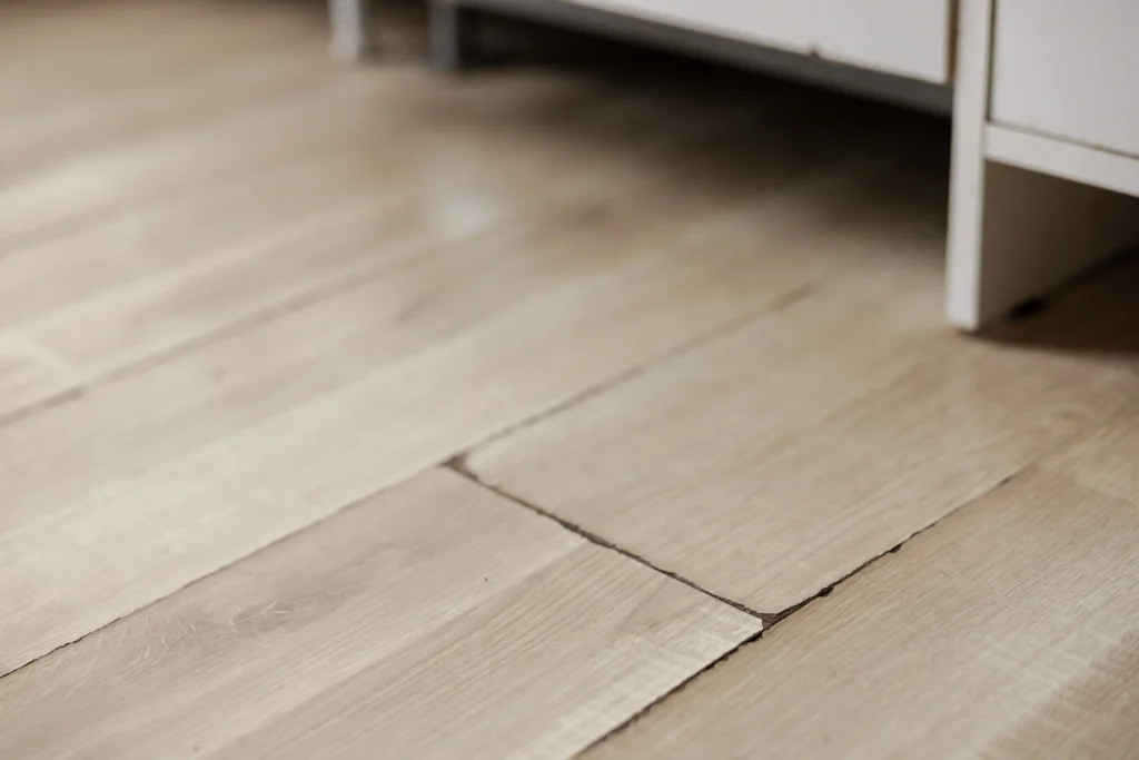 Piso laminado de madeira clara com placas levantadas e desalinhadas próximo a um móvel branco.