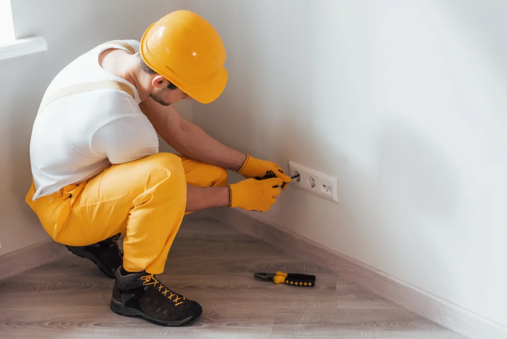 Eletricista residencial utilizando equipamentos de proteção individual e ferramentas para realizar manutenção em uma tomada elétrica na parede de uma residência.